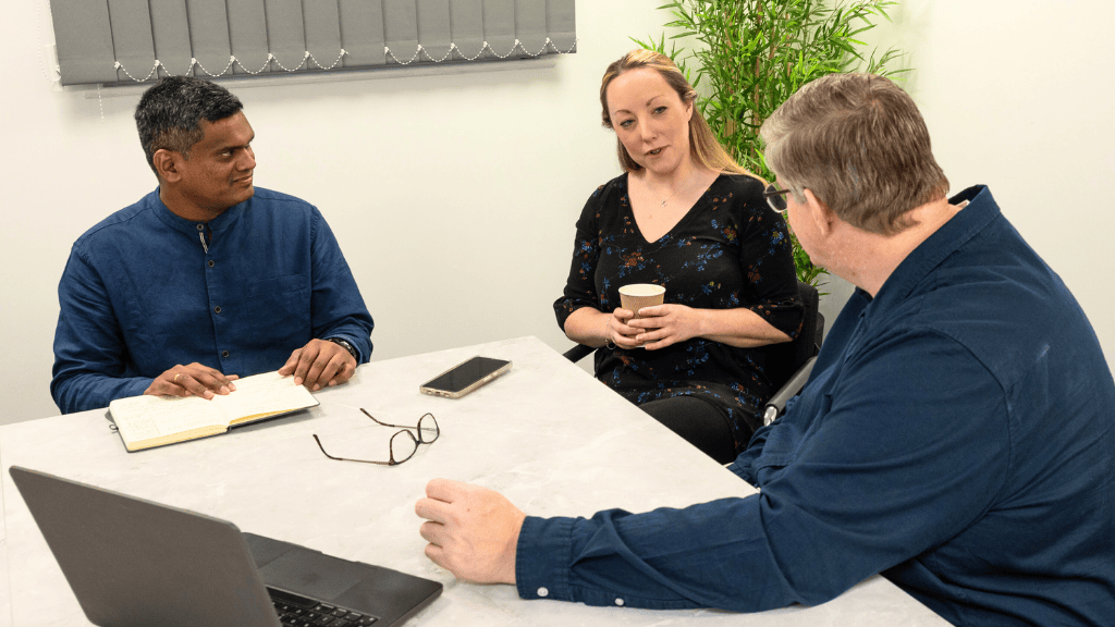 Three people in a business meeting, sitting around a table with a laptop, notebook, and coffee, discussing IT support options
