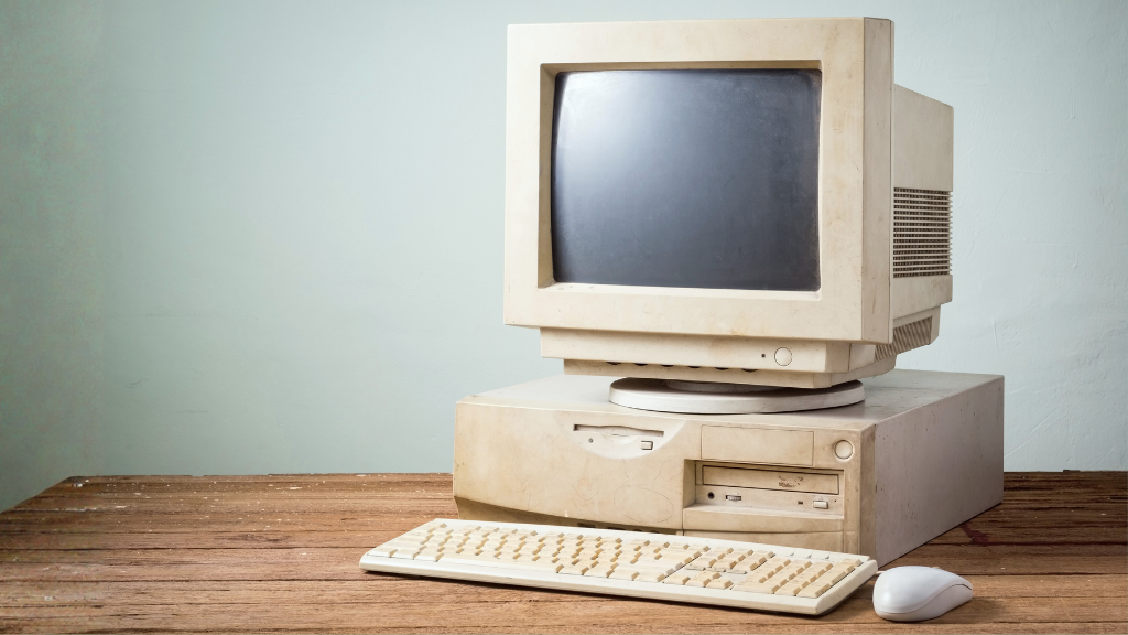 Old and outdated IT system sat on a wooden desk with keyboard and mouse with a light teal/grey wall behind