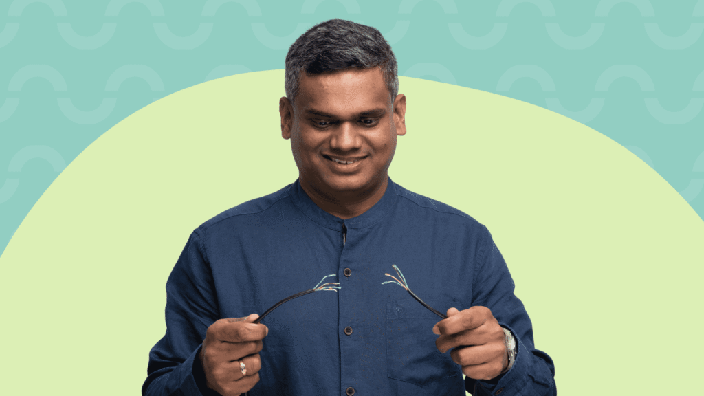 Smiling man in a blue shirt holds his eyeglasses in front of a teal and pale green abstract background.