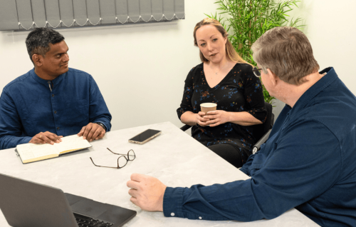 Three people in a business meeting, sitting around a table with a laptop, notebook, and coffee, discussing IT support options