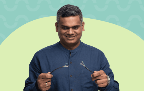 Smiling man in a blue shirt holds his eyeglasses in front of a teal and pale green abstract background.